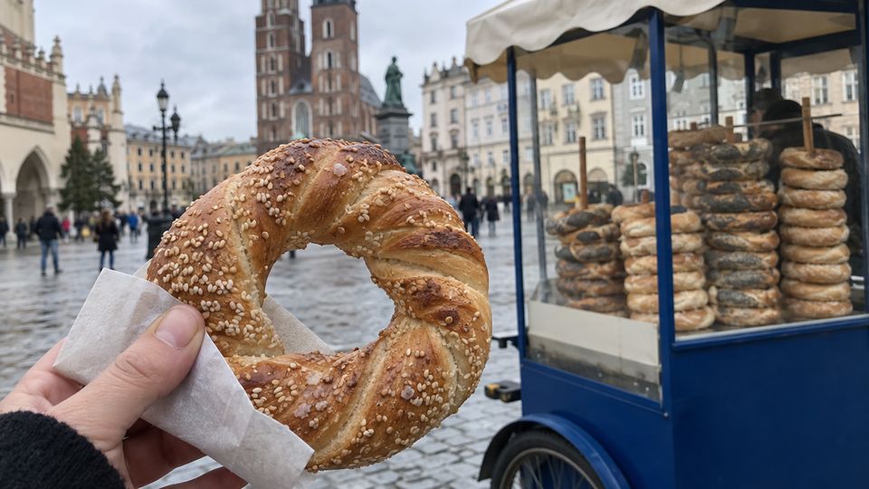A sesame obwarzanek held up in front of a blue obwarzanek cart on Kraków's Main Square
