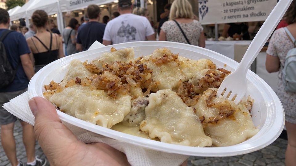 A plastic plate of boiled pierogi topped with fried onion, held up in front of festival stalls and a queue of people on a cobbled square